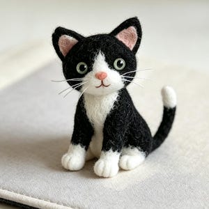 Puede incluir: Una figura de gato de lana fieltrada con pelaje negro y blanco, nariz rosa y ojos verdes. El gato está en posición sentada con la cola ligeramente curvada hacia un lado, mostrando un pecho blanco, patas blancas y una mancha blanca.