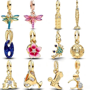 May include: A collection of twelve gold-toned metal charms featuring various designs including a dragonfly, a clock tower, a fish, a blue oval link, a pink flower, a crab, a soccer ball, a castle, a trophy, a cowboy boot, and mouse ears.