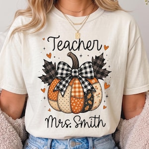 Puede incluir: Una camiseta de manga corta color crema con un gráfico de una calabaza de patchwork con un lazo de cuadros blanco y negro, flanqueada por hojas oscuras. El texto dice Teacher sobre la calabaza y Mrs. Smith debajo en letra negra.