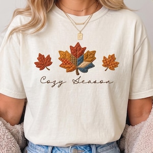 Puede incluir: Una camiseta de manga corta color blanco roto con un gráfico de tres hojas de arce en tonos naranja, amarillo y azul con un patrón acolchado. El texto "Cozy Season" está impreso en letra cursiva marrón debajo de las hojas.