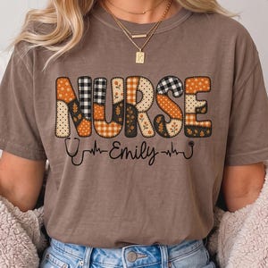 Puede incluir: Una camiseta marrón de manga corta con la palabra NURSE en letras grandes y estampadas con diseños de cuadros, lunares y flores. Debajo del texto, el nombre Emily está escrito en cursiva, flanqueado por un estetoscopio y una línea de pulso.