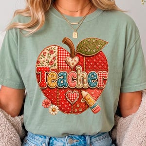 Puede incluir: Una camiseta de manga corta color verde salvia con un gráfico grande de una manzana de patchwork. El diseño de la manzana incluye patrones rojos, blancos y florales, dos corazones pequeños, un lápiz y la palabra Teacher en letras coloridas.