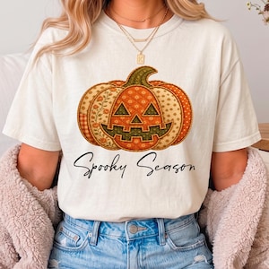 Puede incluir: Una camiseta de manga corta color blanco roto con un gráfico central grande de una calabaza con un patrón estilo patchwork en tonos naranja, verde y crema. Debajo de la calabaza, el texto Spooky Season está impreso en letra cursiva negra.