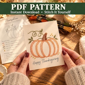 May include: A white card featuring an embroidered pumpkin design with orange thread, green leaves, and gold beads. The text Happy Thanksgiving is printed in black below the pumpkin. A paper pattern sheet with instructions is visible in the background.