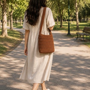 May include: A beige midi dress with short puff sleeves and a round neckline, paired with a brown crocheted shoulder bag featuring a horizontal ribbed texture and a wide strap. The dress has a loose, flowing silhouette and a gathered waistline.