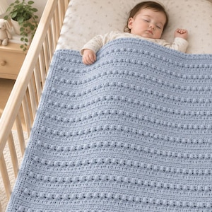 Può includere: Una coperta per neonati all'uncinetto color azzurro con un motivo testurizzato a righe orizzontali di punti nocciolina in rilievo. La coperta rettangolare presenta un bordo semplice ed è adagiata su un neonato che dorme in una culla in legno.