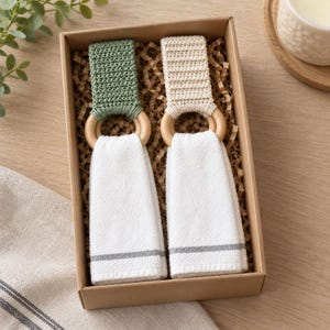 Op de afbeelding: Twee hangende keukendoeken in een kartonnen geschenkdoos. Elke handdoek heeft een witte stoffen basis met een dunne grijze streep, bevestigd aan een ronde houten ring. De bovenste delen zijn gehaakt, één in saliegroen en één in crème.
