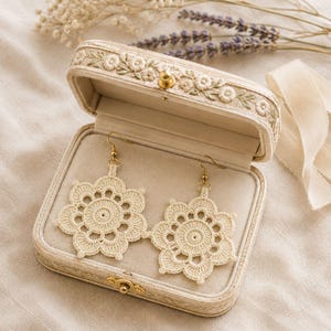 May include: A pair of cream-colored crochet earrings featuring a circular floral pattern with scalloped edges and gold-toned metal hooks. The earrings are displayed inside a rectangular, fabric-covered jewelry box with embroidered floral details.