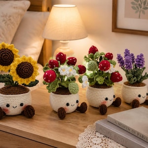 以下が含まれることがあります： 笑顔の顔が刺繍された4つの編みぐるみの鉢植え。1つ目は黄色いヒマワリ、2つ目と3つ目は白い花が咲いた赤いイチゴ、4つ目は紫色のラベンダーです。それぞれの植物はクリーム色の編みぐるみの鉢に入っています。