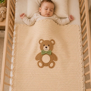 May include: A cream-colored crocheted baby blanket featuring a scalloped edge and a central brown teddy bear appliqué. The bear has a light tan belly, a dark brown nose, and a small green bow tie. The blanket is rectangular with a textured stitch.