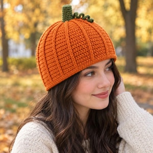May include: A bright orange crochet beanie designed to resemble a pumpkin, featuring vertical ribbed texture, a solid green stem at the top, and a small green curly vine accent. The hat is constructed with a dense stitch pattern throughout.