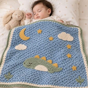 May include: A blue crochet baby blanket featuring a green dinosaur applique, a yellow crescent moon, yellow stars, and white clouds. The blanket has a cream and green border with small green leaf appliques at the bottom corners.