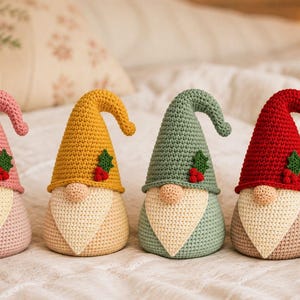 May include: Four crocheted gnome figurines arranged in a row. Each gnome features a tall, pointed hat in pink, yellow, sage green, or red, adorned with a small green holly leaf and three red berries. They have beige noses and cream-colored beards.