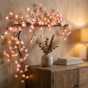 Op de afbeelding: Een decoratieve wandlamp in de vorm van een tak met talloze kleine, oplichtende roze kersenbloesems. De donkerbruine tak loopt horizontaal en buigt naar beneden, verlicht door warme ledlampjes in elke bloemvormige lampenkap.