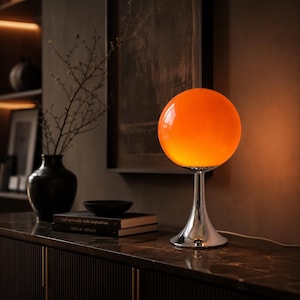 Peut inclure: Une lampe de table composée d'un abat-jour sphérique en verre orange vif monté sur une base en chrome poli avec un pied incurvé en forme de trompette. La lampe diffuse une lueur chaude et douce, soulignant la surface lisse du verre et du métal.