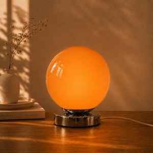 Könnte beinhalten: Eine kugelförmige Tischleuchte aus orangefarbenem Glas mit glatter, glänzender Oberfläche. Die Leuchte verfügt über einen runden, reflektierenden Sockel aus chromfarbenem Metall und ein weißes Stromkabel, das seitlich herausführt.