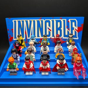 Könnte beinhalten: Ein blauer, abgestufter Präsentationsständer mit dem Wort INVINCIBLE in großen weißen Buchstaben und roten Spritzern. Der Ständer enthält neunzehn kleine Kunststofffiguren mit verschiedenen bunten Outfits, Umhängen und Zubehör, die in Reihen angeordnet sind.