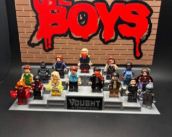 May include: A display stand featuring fifteen miniature figures arranged on tiered platforms. The background is a brick-patterned sign with the red text "THE BOYS". The base of the stand features the "VOUGHT INTERNATIONAL" logo in black and white.