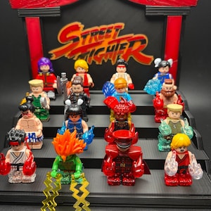 Puede incluir: Una colección de quince minifiguras con temática de Street Fighter dispuestas en un expositor negro escalonado con un encabezado rojo y negro que muestra el logotipo de Street Fighter. Cada figura tiene efectos de energía de plástico translúcido.