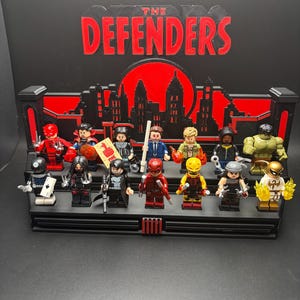 May include: A black display stand featuring the red text THE DEFENDERS and a city skyline silhouette. It holds fourteen miniature figures, including characters in red, black, blue, and green outfits, some holding accessories like weapons and cards.
