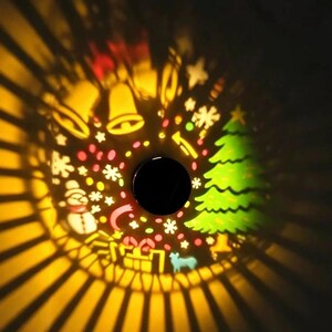 May include: A circular light projection displaying festive holiday icons including a green Christmas tree, a white snowman, yellow bells, gift boxes, and various snowflakes, all surrounded by radiating beams of warm yellow light.