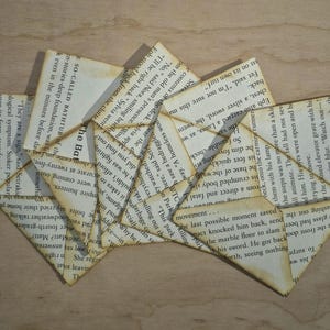 May include: A collection of triangular page corner bookmarks crafted from vintage book pages with visible printed text. The paper features a light tan, aged patina and is folded into geometric shapes with sharp, pointed edges and visible creases.