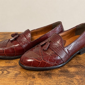 May include: A pair of dark burgundy loafers featuring a combination of smooth and textured leather panels with a crocodile-skin pattern. Each shoe is adorned with a decorative leather tassel on the vamp and has a low, stacked heel.