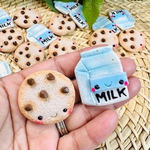 May include: A round tan cookie with brown chocolate chips and a small smiling face, alongside a light blue milk carton with a smiling face and the word MILK printed in black. Both items feature pink cheeks and are crafted with a textured finish.