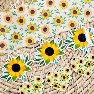 May include: A collection of sunflower-themed craft supplies including a long strip of cream-coloured ribbon with a repeating sunflower pattern and several die-cut stickers featuring individual sunflowers and heart-shaped sunflower prints.