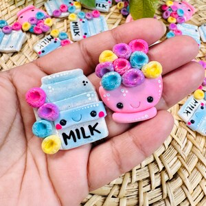 Op de afbeelding: Twee kleine kleibedels bestaande uit een lichtblauw melkpak met het woord MILK in het zwart en een roze kom gevuld met kleurrijke ontbijtgranen. Beide items hebben lachende gezichtjes met zwarte ogen en zijn voorzien van glitters.