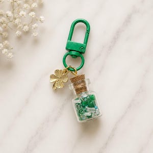 May include: A green metal lobster clasp keychain featuring a gold-toned four-leaf clover charm and a small clear glass bottle filled with assorted green, white, and translucent beads, sealed with a natural brown cork stopper.
