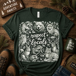 Può includere: Una t-shirt verde scuro con una stampa grafica bianca di vari animali della foresta, tra cui un procione, un orso, una volpe, un gufo, un coniglio, un castoro, un cervo e una lince, con il testo "respect the locals" al centro.