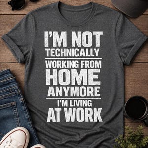 May include: A dark charcoal gray short-sleeved t-shirt featuring white distressed-style text that reads: I'M NOT TECHNICALLY WORKING FROM HOME ANYMORE I'M LIVING AT WORK. The shirt has a crew neckline and a standard fit with rolled sleeves.
