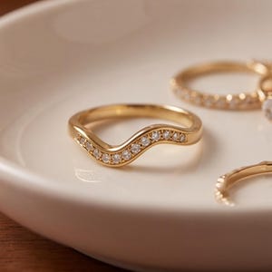 May include: A gold wave-shaped ring featuring a row of small, clear, round-cut stones set along the top curve. The ring is placed inside a smooth, white, shallow ceramic dish alongside other gold rings with similar stone settings.