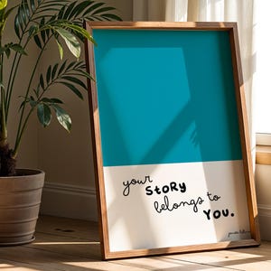 May include: A framed art print with a teal and white color block design. The artwork features the handwritten text "your story belongs to YOU." The frame is made of wood and is leaning against a wall near a window and a potted plant.