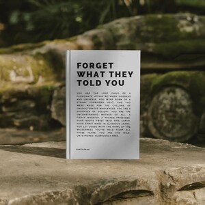 May include: A hardcover book with the title "FORGET WHAT THEY TOLD YOU" in bold black letters. The book is white and features black text on the cover and inside. The book is resting on a stone surface.