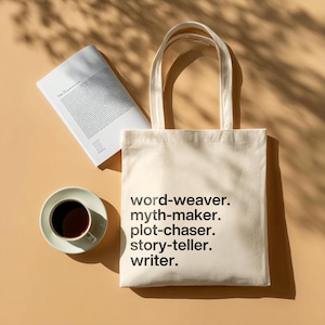 May include: Cream-colored tote bag with black text: "word-weaver. myth-maker. plot-chaser. story-teller. writer." Beside it, a book and a cup of coffee on a saucer. The items are on a light brown surface with shadows.