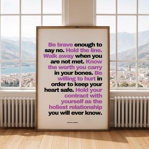 Purple and black boundary print in a thin dark wood frame standing on a pale wood floor in front of a tall window with a view of mountains, a lake, and a hillside town, between two white radiators. Bright, clear daylight fills the room. A calm, open setting for counselor office decor in a practice with big windows, where clients can sit, breathe, and talk.