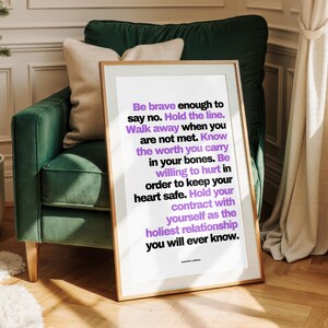 Purple and black boundary print in a thin light wood frame with a white border leaning against a dark green velvet armchair with a cream pillow. Paneled cream walls, a white radiator, linen curtains, a small potted tree, and a herringbone floor sit around it. The purple pops against the deep green. A personal power piece for a reading corner, and a courage print for a woman learning to hold the line.