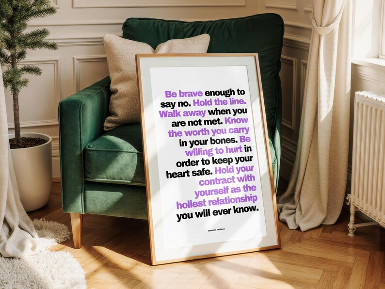 Purple and black boundary print in a thin light wood frame with a white border leaning against a dark green velvet armchair with a cream pillow. Paneled cream walls, a white radiator, linen curtains, a small potted tree, and a herringbone floor sit around it. The purple pops against the deep green. A personal power piece for a reading corner, and a courage print for a woman learning to hold the line.
