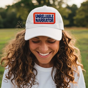 May include: White baseball cap with the text "UNRELIABLE NARRATOR" in blue letters inside a red and white rectangle. The cap is worn by a person with long, curly brown hair.