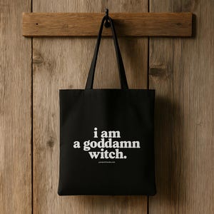 以下が含まれることがあります： 「i am a goddamn witch.」の白文字がプリントされた黒色のトートバッグ。バッグは黒いフックに掛けられ、木の板に吊り下げられています。背景は木の壁です。