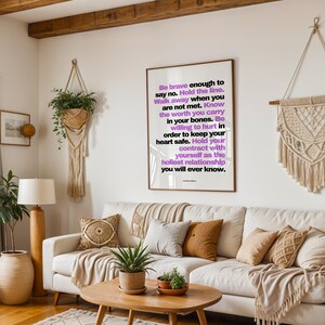 Purple and black boundary print in a light wood frame hanging above a cream sofa with tan and patterned pillows, between a trailing plant in a macrame hanger and a large cream macrame wall hanging. A wood beam ceiling, a floor lamp, potted plants, and a round wood coffee table fill the boho living room. Shown at eye level in a shared space. A manifesto print for a home where everyone is learning healthy limits.