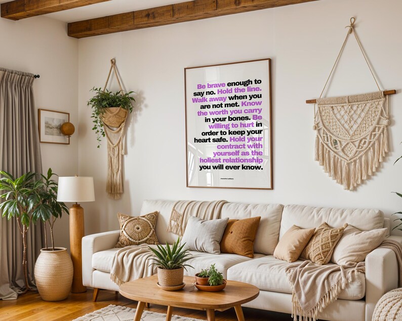 Purple and black boundary print in a light wood frame hanging above a cream sofa with tan and patterned pillows, between a trailing plant in a macrame hanger and a large cream macrame wall hanging. A wood beam ceiling, a floor lamp, potted plants, and a round wood coffee table fill the boho living room. Shown at eye level in a shared space. A manifesto print for a home where everyone is learning healthy limits.