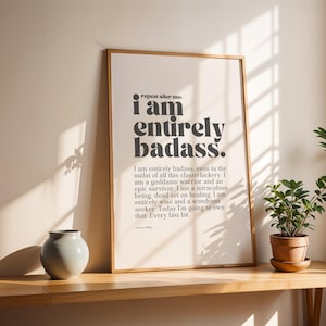 May include: A framed print with the text "I am entirely badass." in a bold, black font. The print is in a wooden frame and is displayed on a wooden table. A small, light blue vase and a potted plant are also on the table.