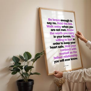 A person in a cream long sleeve top hangs the framed purple and black boundary print on a plain cream wall, holding the light wood frame by both sides, beside a dark green plant in a black pot and a small vase of dried stems. Shows the poster at eye level as it goes up. A clear, readable choice of therapy office art for a counselor, coach, or social worker setting up a new room.
