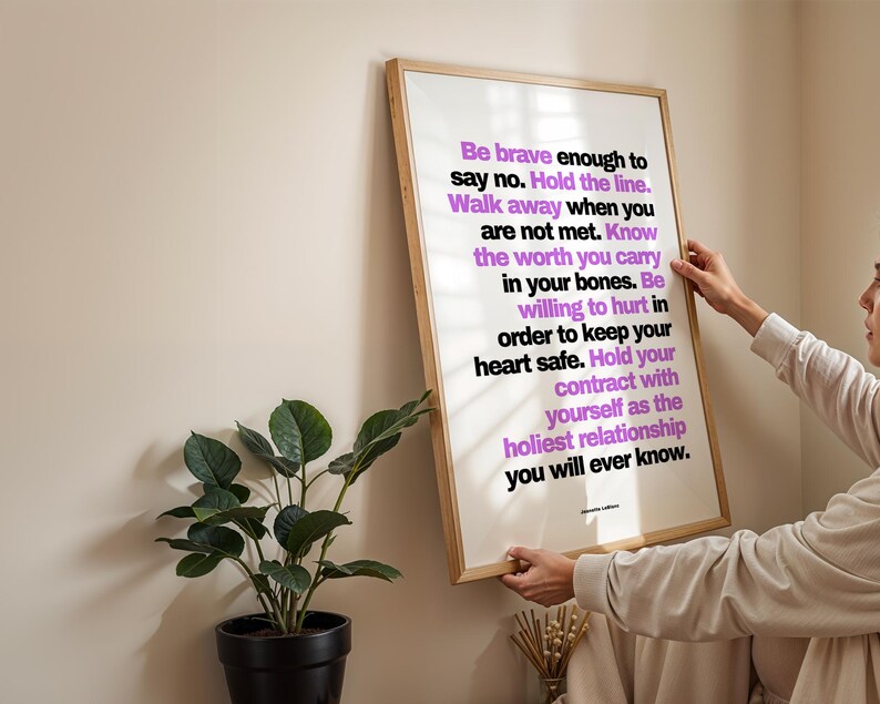 A person in a cream long sleeve top hangs the framed purple and black boundary print on a plain cream wall, holding the light wood frame by both sides, beside a dark green plant in a black pot and a small vase of dried stems. Shows the poster at eye level as it goes up. A clear, readable choice of therapy office art for a counselor, coach, or social worker setting up a new room.