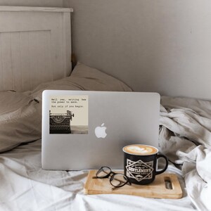 Cozy MacBook setup with a sepia-toned sticker reading “Hell yes, writing has the power to save. But only if you begin.” A Jeanette LeBlanc quote offering storytelling encouragement and daily writing motivation.