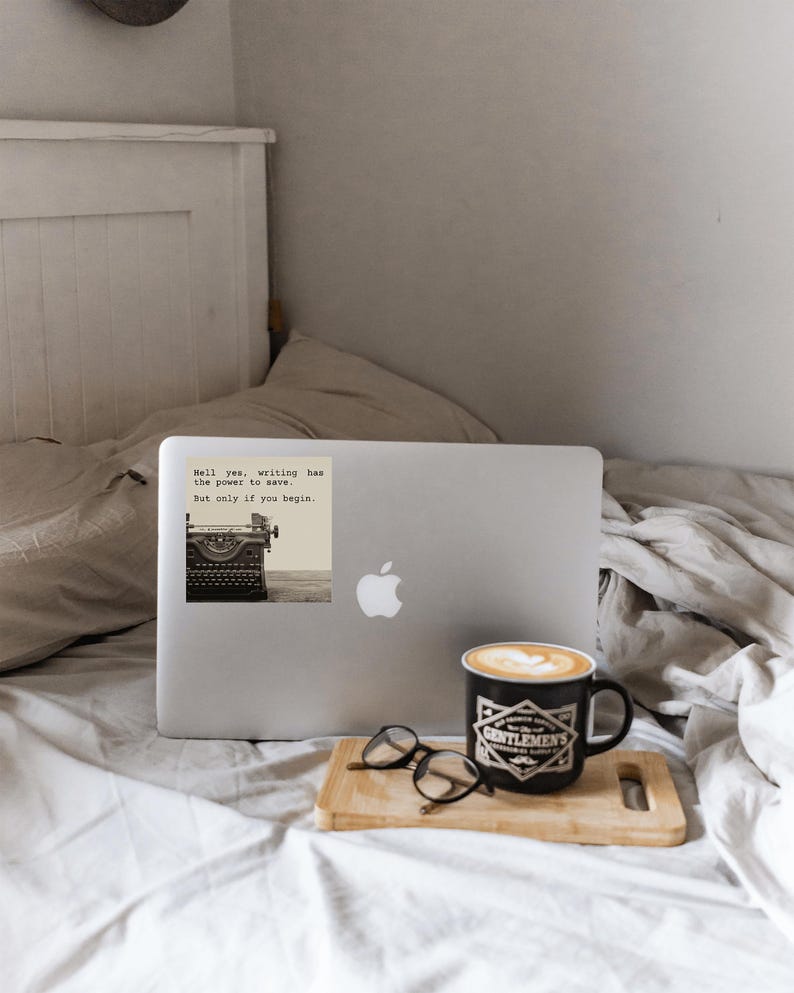 Cozy MacBook setup with a sepia-toned sticker reading “Hell yes, writing has the power to save. But only if you begin.” A Jeanette LeBlanc quote offering storytelling encouragement and daily writing motivation.