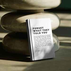 May include: A white book with the text "FORGET WHAT THEY TOLD YOU" in bold black letters. The book is propped up against a stack of three off-white, rounded, stacked objects. The book's text continues with inspirational prose.